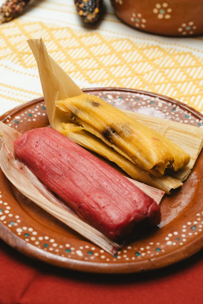 Two tamales, one pink and one yellow, served on a traditional clay plate with corn husks.