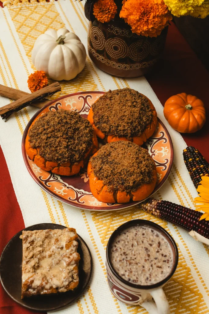 Autumn-themed setup with pumpkin conchas, a slice of dessert, and a mug of champurrado on a festive table with marigolds and mini pumpkins.