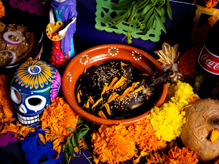 Traditional Día de Muertos altar decorated with marigold flowers, colorful papel picado, a painted sugar skull, a clay plate of mole with chicken topped with sesame seeds and marigold petals, a pan de muerto bread, a figurine of a painter, and a glass bottle of Coca-Cola.