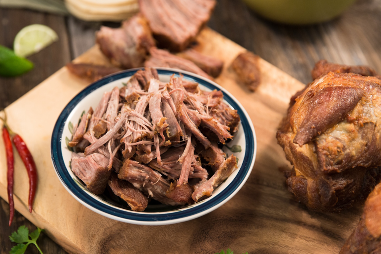 Bowl of shredded carnitas on a cutting board with a ingredients on the side.