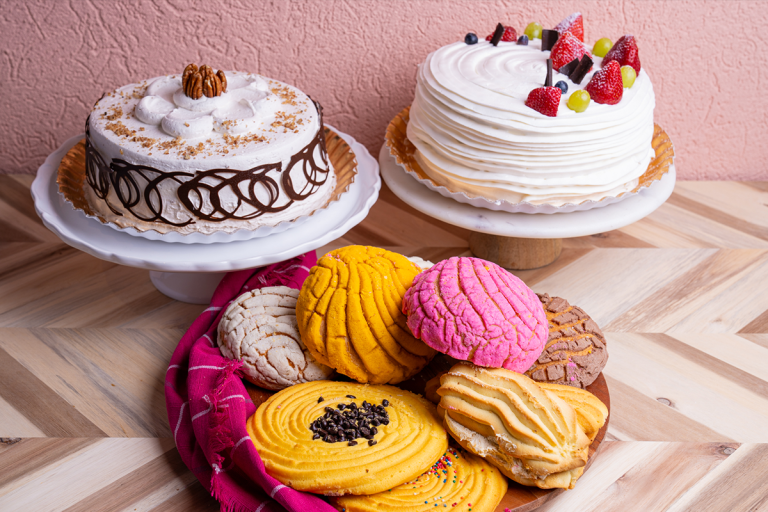 Beauty shot of white cakes and pan dulce on a wood table with a pink background.