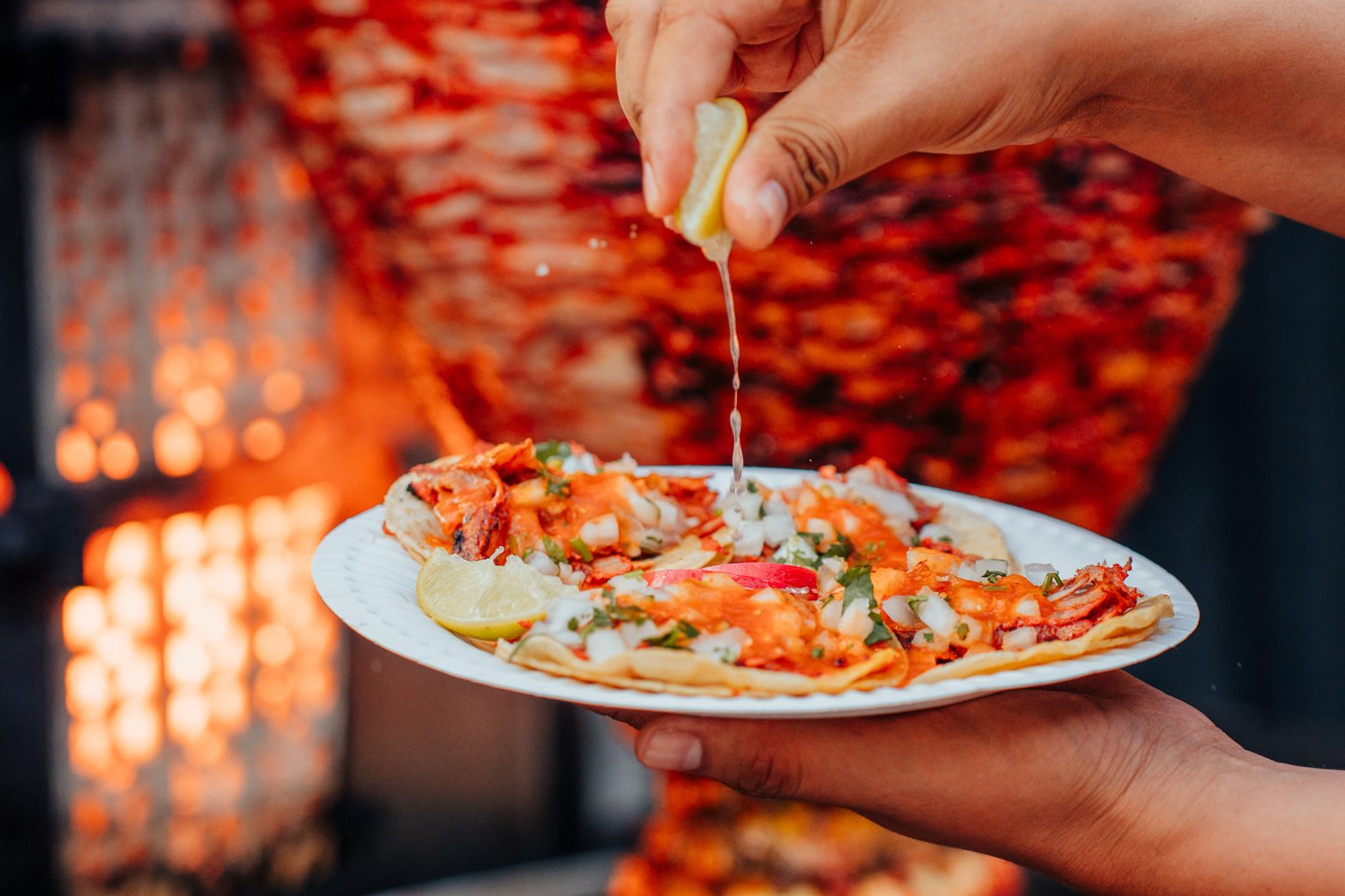 Tacos Guichos with trompo in the background and a person squeezing lime on the taco